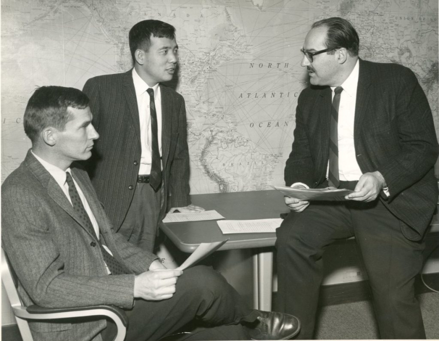 Black and white photo of Joseph Smagorinsky, founding director of GFDL, with Kirk Bryan (seated on left in chair) and his research partner, Syukuro “Suki” Manabe (standing), discussing a paper they were writing on the coupled atmosphere-ocean model in 1969.