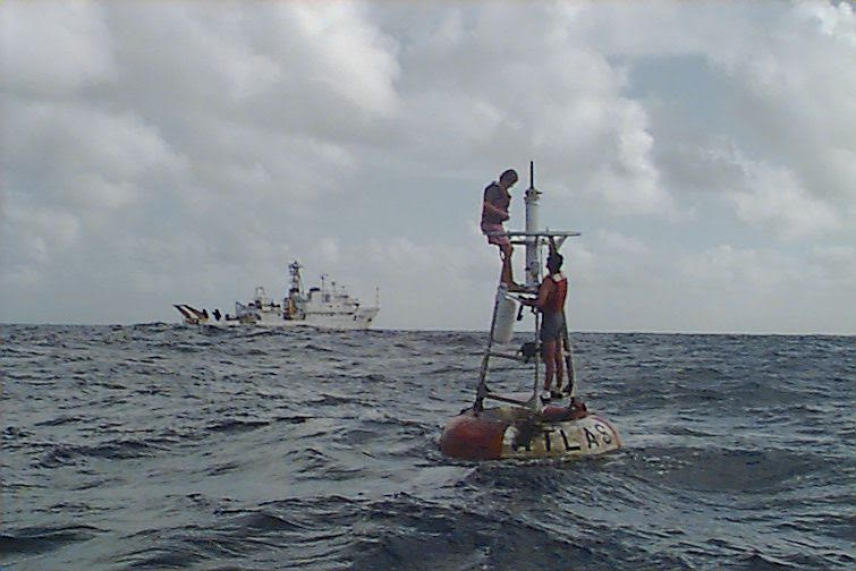 Photo of two men working to repair a wind sensor on a Tropical Atmosphere Ocean buoy moored near Hawaii in 1996. In the background, the NOAA ship Ka'imimoana.