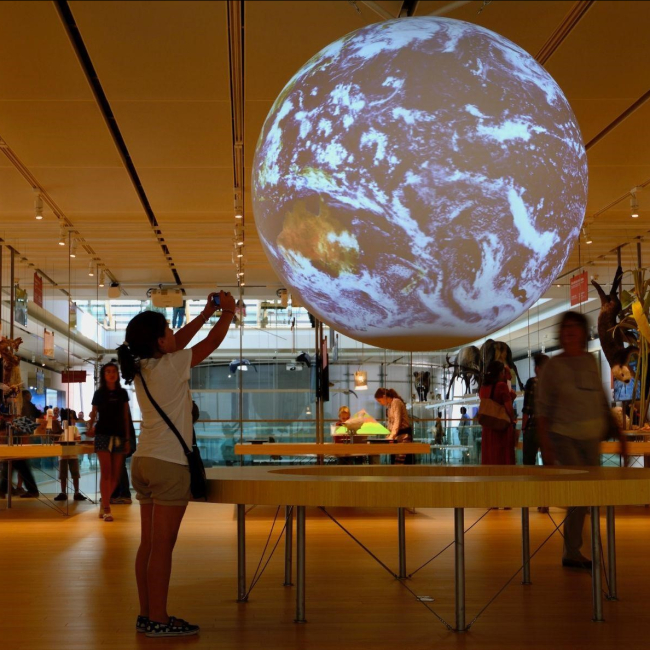 A young girl takes a photo of a 6-foot animated globe showing a color view of the Earth with clouds of the globe at a museum exhibit.