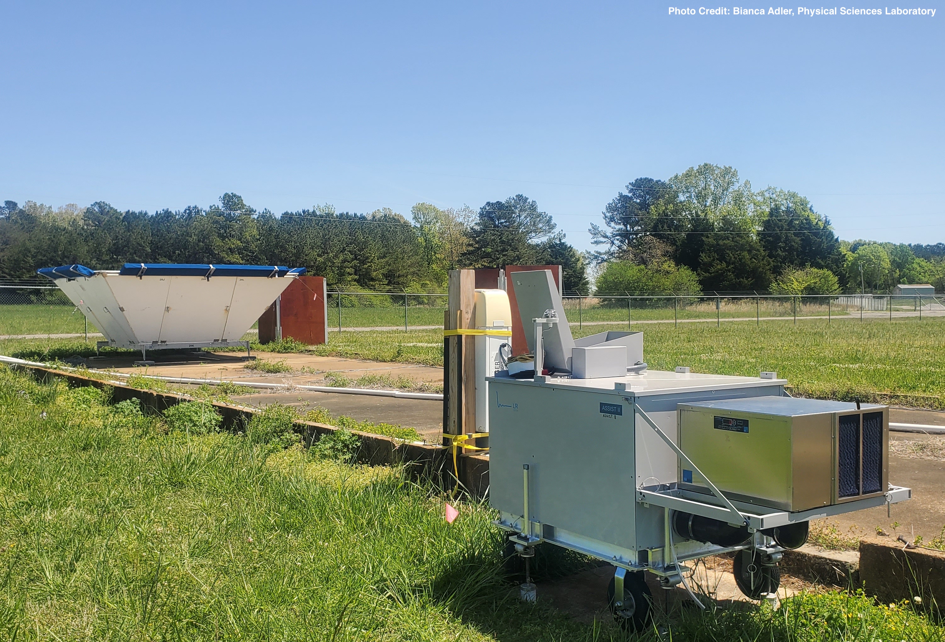 An infrared spectrometer and ceilometer sit in the foreground with a trapezoidal radar wind profiler in the background, in a grassy field in Courtland, Alabama.