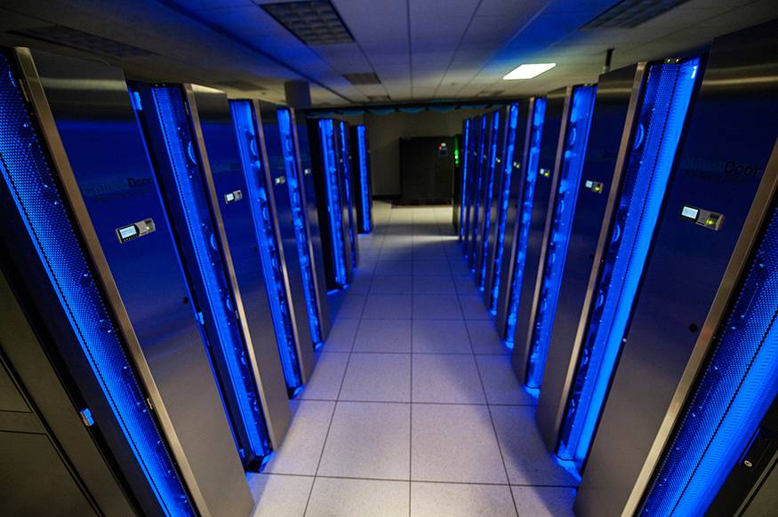 Image is of the Mississippi State University Orion supercomputer located in Starkville, MS. Orion is a Dell supercomputer with 72,000 Skylake Processors and consists of multiple black with blue lighting cabinets (16 are shown in the image). The racks of hardware components are stacked vertically to save space,  allow for ease of connecting the many nodes and cores of the supercomputer, and improve efficiency in cooling. The racks are also labeled with Chilled Door Rack Cooling system and have a status