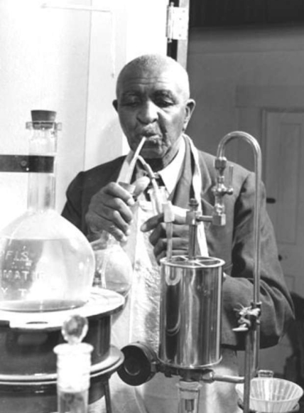 Black and white photo of George Washington Carver working in his laboratory. He stands among laboratory devices holding a flask with a tube inserted into it. The other end of the tube is in his mouth.