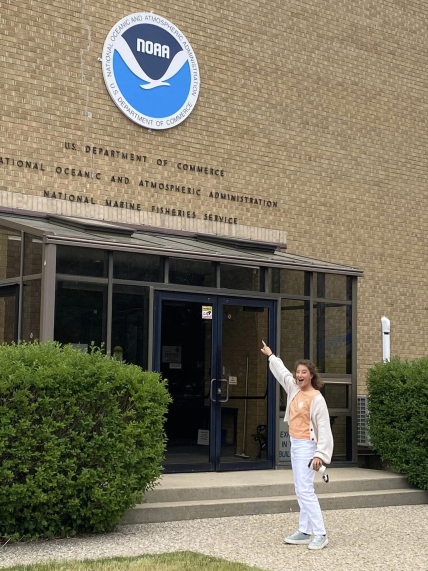 Allie stands outside of a large brick building and points to a NOAA logo and text above the door that reads "U.S. Department of Commerce, National Oceanic and Atmospheric Administration, National Marine Fisheries Service.