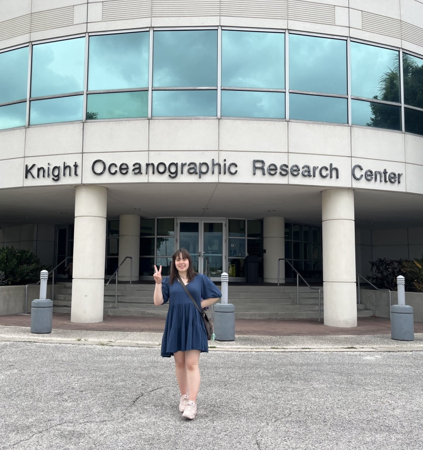 Anna poses in front of a building and smiles to the camera, making a peace sign. The concrete building reads "Knight Oceanographic Research Center" in large letters. Above that, a line of windows reflect cloudy skies and tropical looking vegetation.