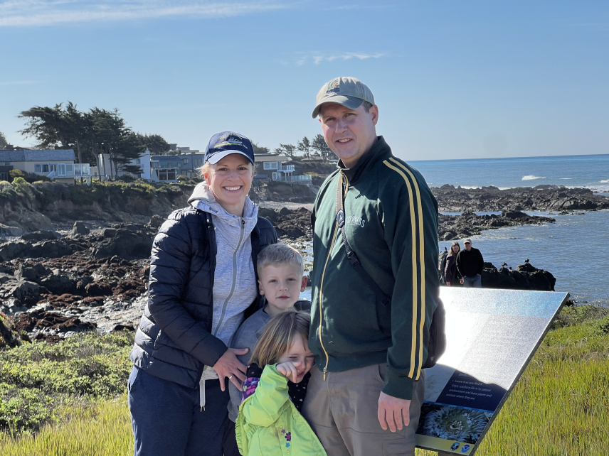 Two adults stand on either side of two children outside on a grassy hiking trail next to a sign. A few houses sitting above the ocean and a rocky shore are visible in the background. 