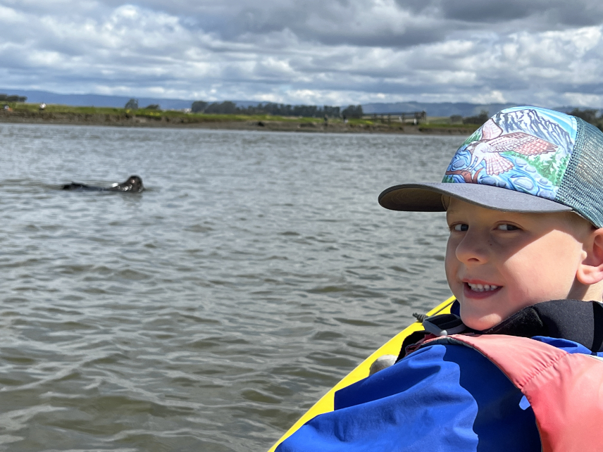 A child with a baseball cap poses inside a kayak near a sea otter swimming at the surface of the water. A green landscape and some mountains are in the background. 