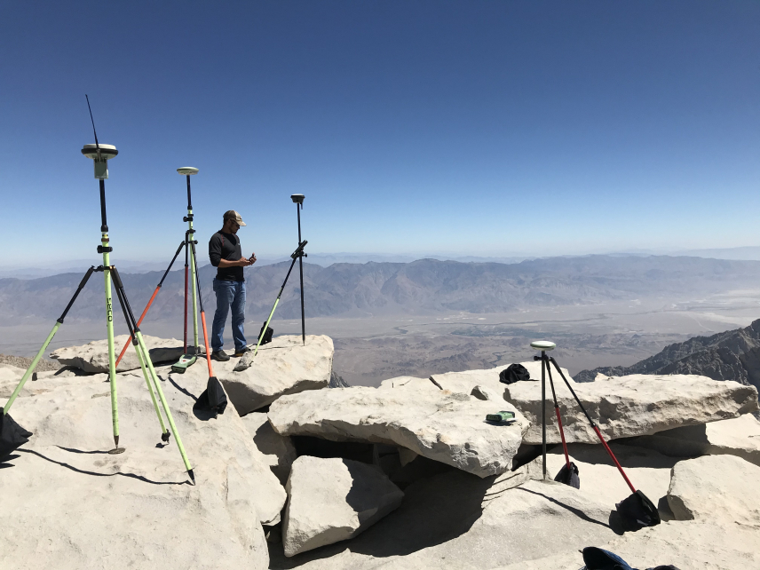 Philip Melcher consults a handheld GPS while standing on a rocky outcropping overlooking a vast mountainous range. He is surrounded by four large tripods holding devices used for making GPS observations, one of which is positioned directly over a small metal disk attached to the rock.