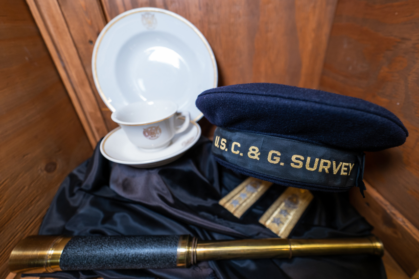 A U.S. Coast & Geodetic Survey (USC&GS) cap sits on a dark wooden background, along with a plate, tea cup, and saucer from the U.S. Coast and Geodetic Survey, as well as a brass telescope. The  black cap is shaped like a beret with a wide band at the bottom. On the band, "U.S. C. & G. SURVEY" is written in gold.