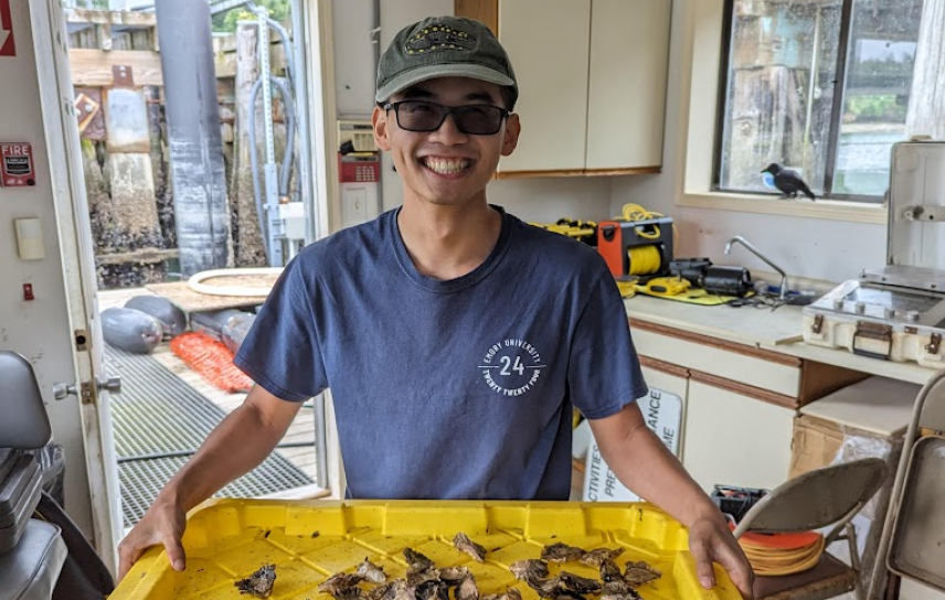 Nick stands in what looks to be a lab for processing field samples. He grins, holding a large tray with oyster shells. His field boots and active transition lenses implied he's just brought them inside.