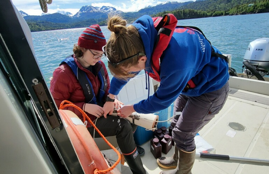 Seraina and Nicole are aboard a small research vessel on what looks to be calm waters. Seraina is manipulating a cylindrical instrument that Nicole holds in her lap. They are dressed for cold weather fieldwork and wear personal floatation devices.