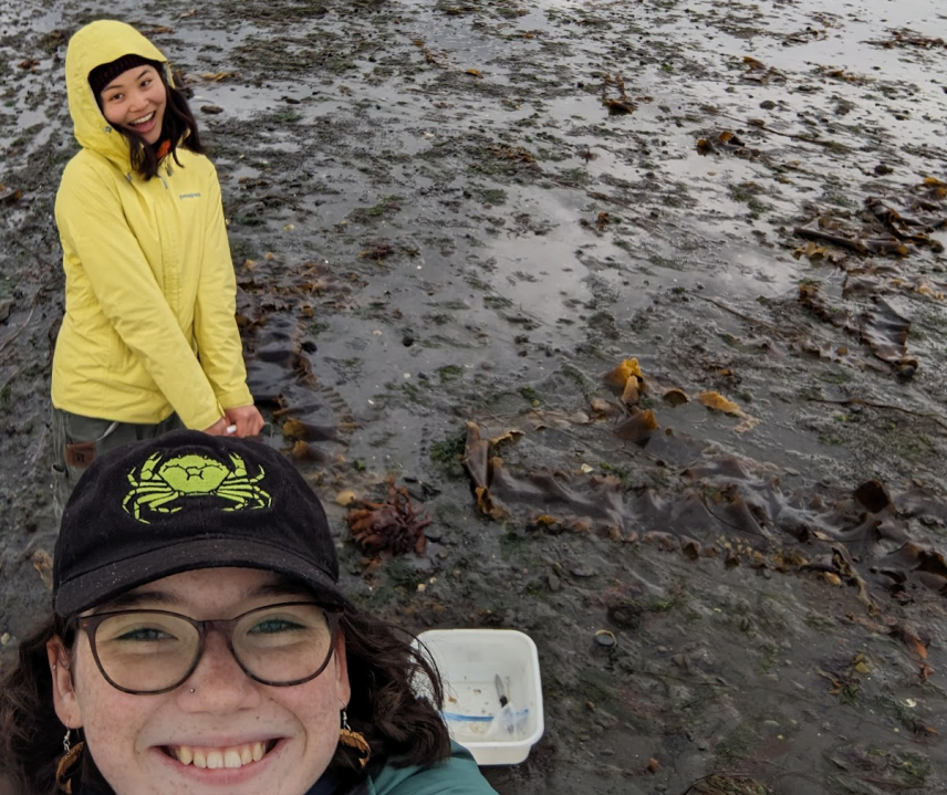 Nicole and Eojin smile for a selfie taken by Nicole. They are standing in a bay where the tide has receded, revealing muddy sediment covered in seaweed and algae.
