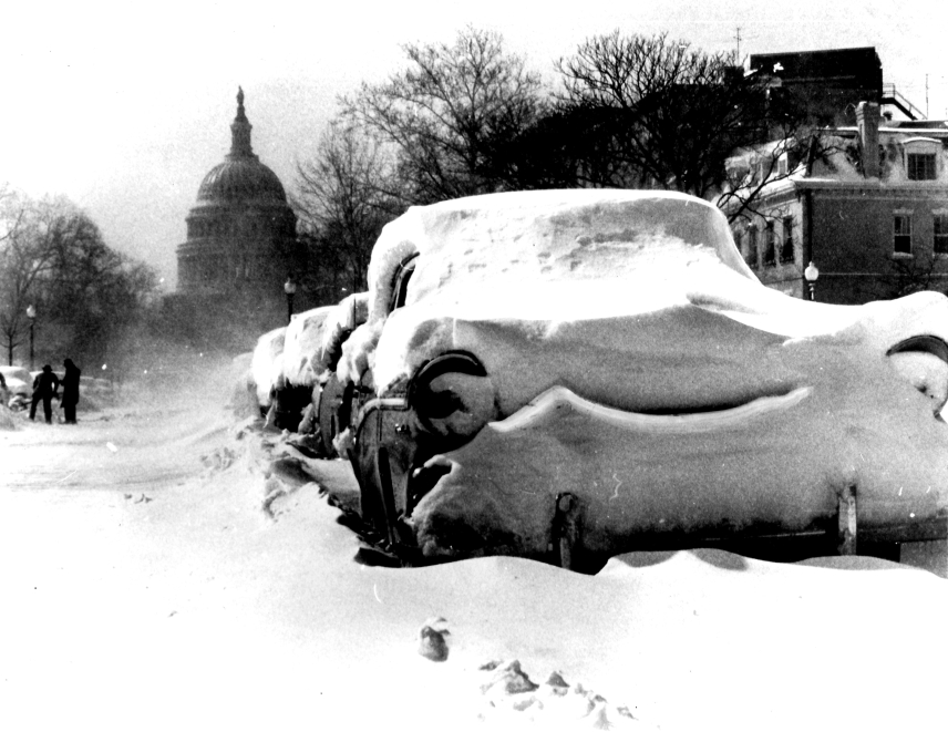 Black and white photo of a line of cars buried in snow with the U.S. Capitol building in the background.
