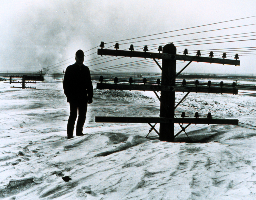 Black and white photo of North Dakota DOT employee, Bill Koch, standing next to the top of a utility pole in North Dakota during a blizzard. March 9, 1966.
