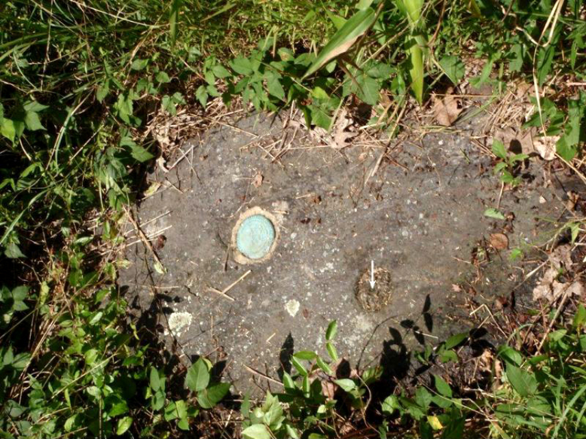Photo of a small cleared area within a grassy border. A white arrow points to Hassler’s original survey marker, which was placed in 1833 and composed of an iron pipe embedded into the bedrock. USGS added the bronze disc on the left, now with a blue/green patina, in 1932.