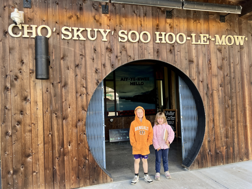 Two children stand in front of a circular entrance to a wood-paneled building. Above the entrance are the Yurok words “Cho’ Skuy’ Soo Hoo-Le’-Mow’,” which means “May You Folks Travel Safely” in English. 