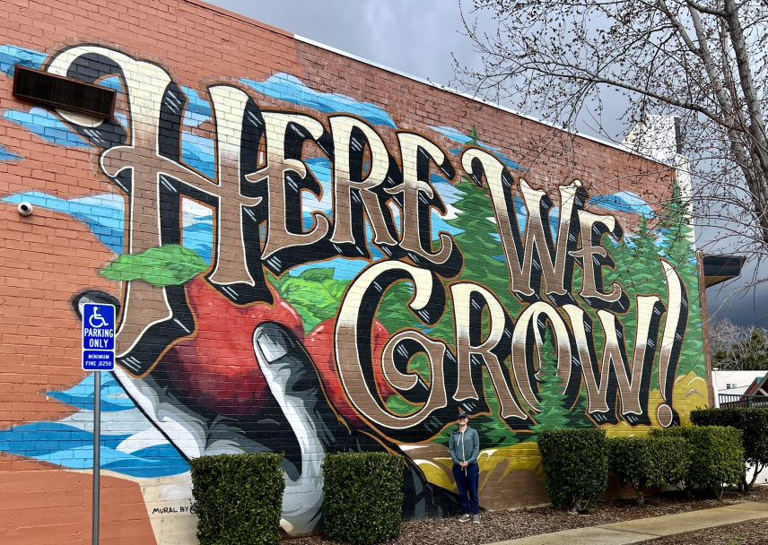 An adult in a sweatshirt and baseball caps stands outside in front of a large mural that reads, “Here We Grow.” The mural contains a hand holding two apples and pine trees and is painted on a brick building.