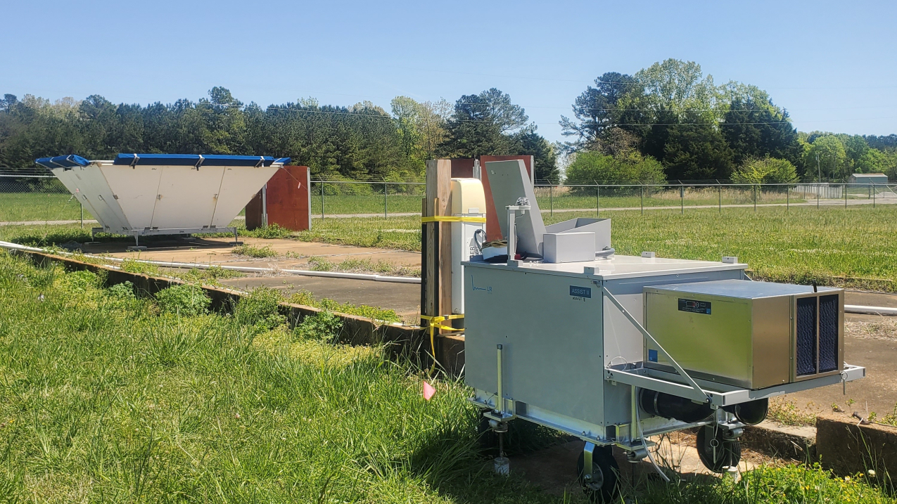 An infrared spectrometer and ceilometer sit in the foreground with a trapezoidal radar wind profiler in the background, in a grassy field in Courtland, Alabama.