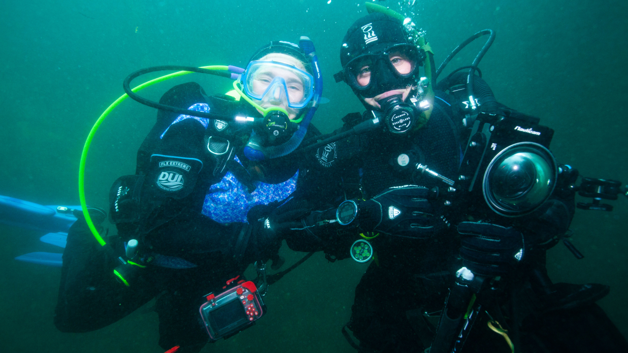 Audrey and Gracia are underwater in full SCUBA gear. They pose for the camera and though their mouths are difficult to see around the scuba mouthpiece, their eyes are clearly smiling. Gracia holds a large, professional-looking underwater camera while Audrey has a smaller underwater camera.
