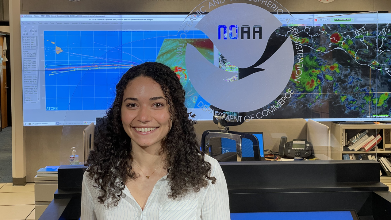 Delián poses  in business casual clothing and smiles for the camera in front of a glass wall with an etched NOAA logo on it. In the background, the words "National Hurricane Center, National Weather Service" are written on the wall above two large monitors showing satellite weather images.