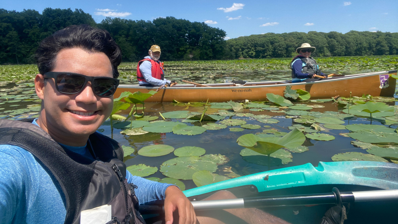 A selfie-style photo taken by Keanu. He smiles in the foreground in his kayak. Two people are in a canoe in the background and smile for the camera. Everyone is wearing protective life vests and sun protection as they float among the lotus and lily pads on a calm sunny day.
