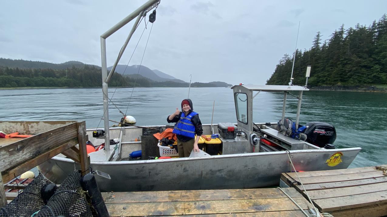 Cassy smiles and gives a thumbs up while standing on a small, docked research vessel that holds assorted sampling gear, and has an a-frame with a winch. She is wearing a hooded windbreaker and life jacket over a pair of waders. The day is overcast and waters are calm, and mountains and an evergreen forest surround the body of water.