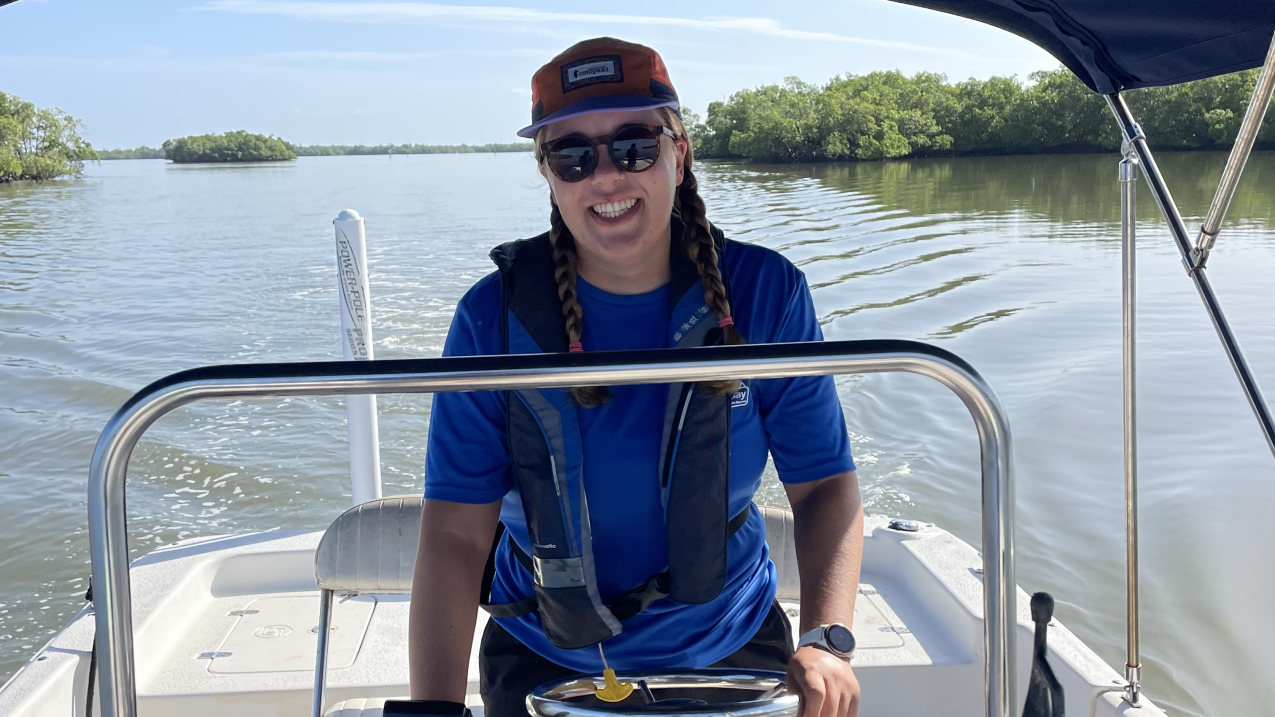 Ellie stands at the helm of a center console boat, driving with a smile on her face in calm inland waters. They are cruising at a slow speed judging by the wake. She wears a personal flotation device and equipment is stored neatly to the sides of the boat.