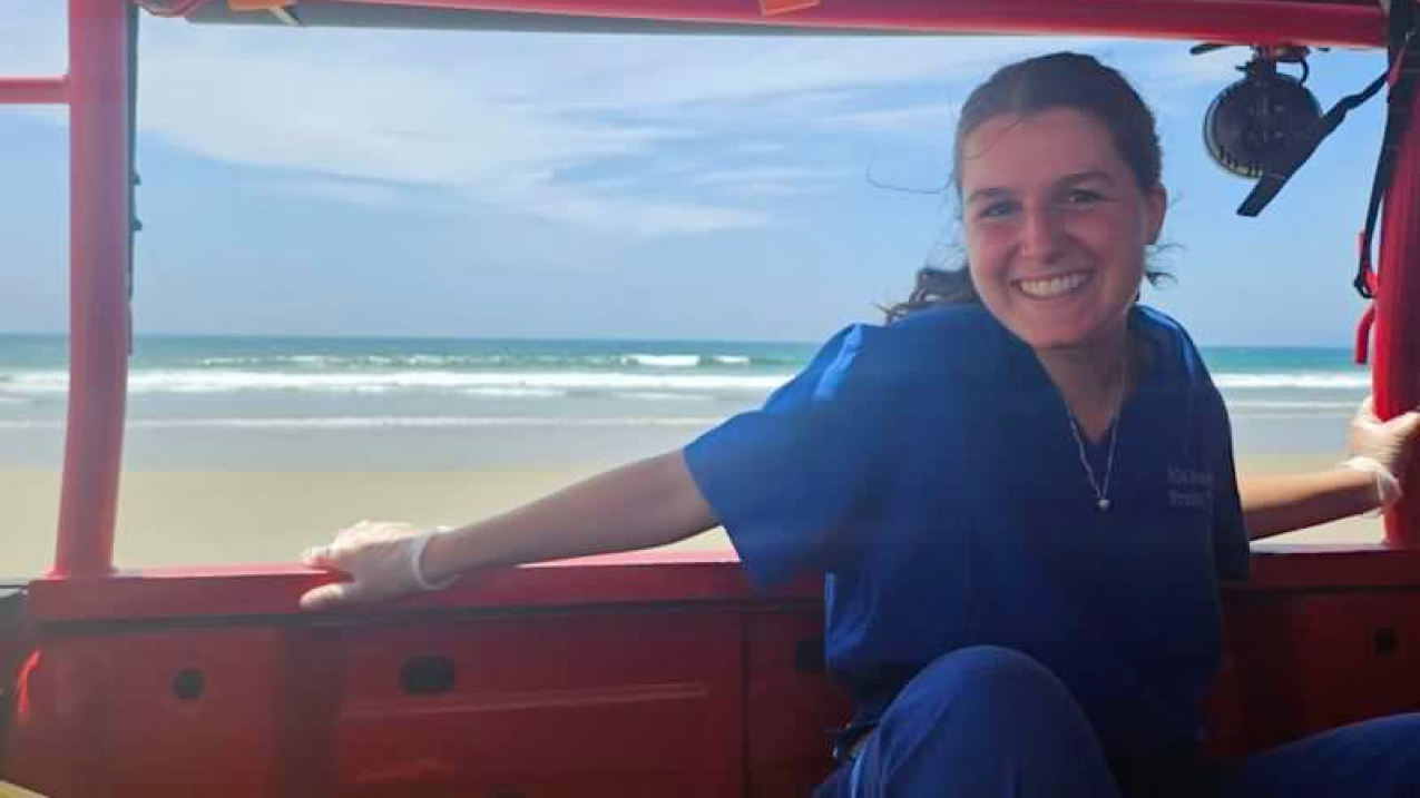 Sarah smiles, sitting in an open-air vehicle, with an empty beach visible through the opening behind her, as though the vehicle is driving on the beach. She is wearing an outfit similar to surgical scrubs and rubber gloves.