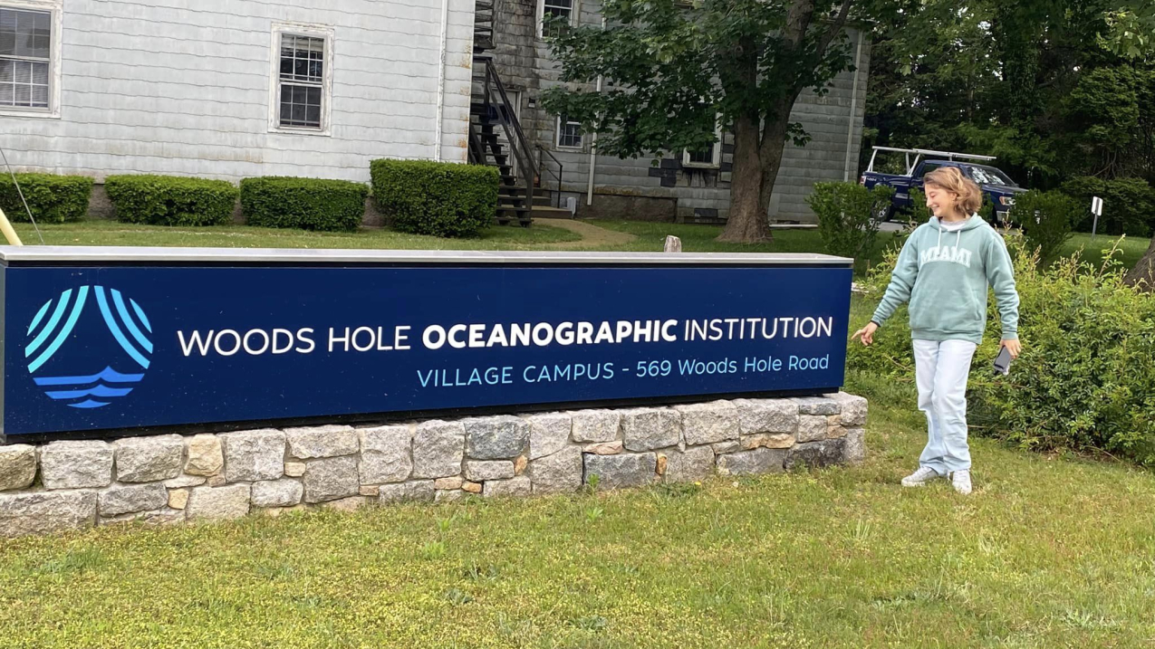 Allie stands outside next to a sign that reads "Woods Hole Oceanographic Institution. Village Campus.
