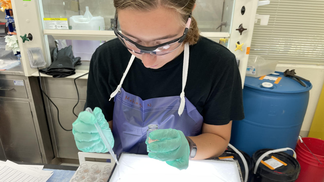 Makayla stands at a lab bench, looking down at a lab tray that looks to contain shallow water. She holds a transfer pipette in one gloved hand and a sampling vial in the other. She wears protective goggles and an apron.