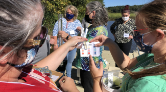 Teachers stand on dock. Photo focuses on 2 teachers holding a vile of water and a water quality card as they compare the color of the water they collected in the vile to the colors on the card.