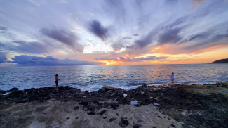 Two students stand on the shore of an island and cast fishing lines into the water. There’s a beautiful orange and blue sunset over the ocean.