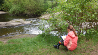 An elementary student sits on the grass along a creek wearing a raincoat and rain boats. She is writing on a clipboard.