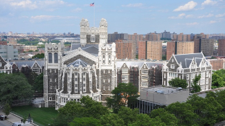 Several buildings in English Gothic style, built with dark stone and white trim. Trees surround the campus in the foreground and a large city sits behind the campus.