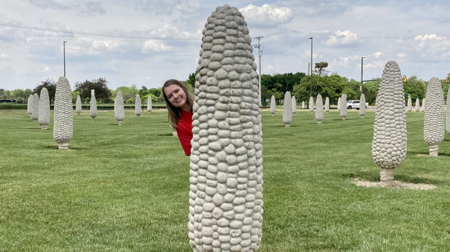 Maggie Beetstra stands behind a large stone sculpture of an ear of corn