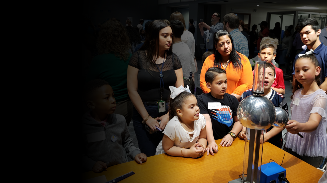 Young children gather around a Van de Graaff machine, a metal structure with a large metal sphere on top, with a metal spherical wand attached. The children watch the machine with faces of awe as they see electricity passing between the machine and the wand.