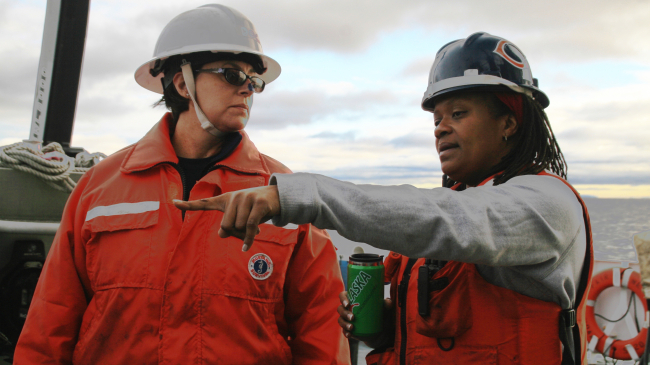 Two people stand on a large ship wearing protective hard hats and high visibility gear. The person on the right is pointing to something off camera. The ocean is visible in the background. 