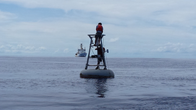 technicians workin on a TAO buoy on the open sea with an approaching ship in the distance
