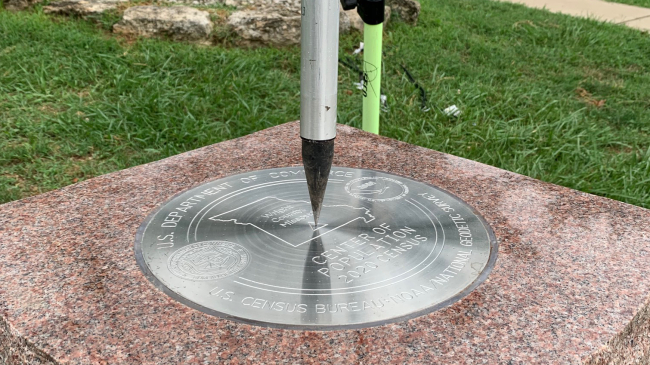 Photo showing The tip of a tripod resting on the center of the 2020 Center of Population Commemorative Survey mark, as part of a GPS survey to determine the precise latitude, longitude, and height of the mark in Hartville, Missouri.