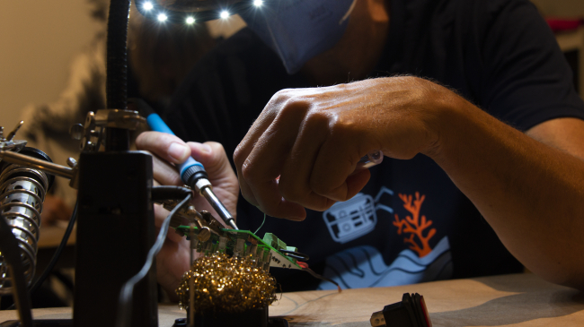 Teacher at Sea alumnus Jeff Miller sits at a workbench, his face mostly obstructed from view by a large magnifying glass ringed with bright lights. A small, square green circuit board is mounted at an angle underneath the light. The focus of the photo is on Jeff’s hands.
