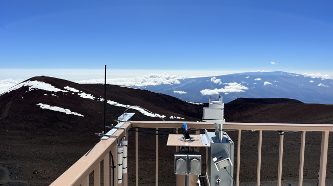A rectangular tube is attached to the banister of a deck on a building. The deck overlooks mountains with some snow cover and a two lane road. Cloud cover is below the deck height, down in the valleys. 