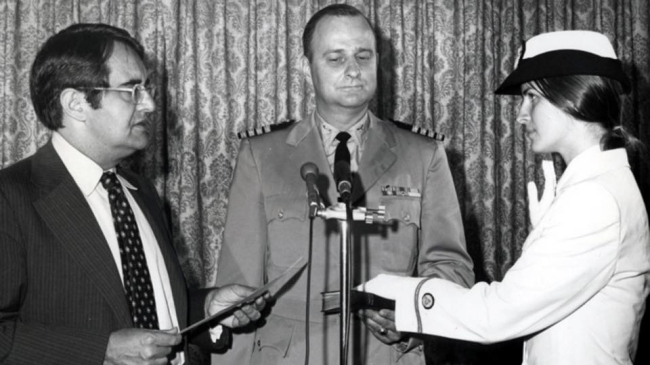 A black and white photo of Ensign Pamela Chelgren being sworn in by Secretary of Commerce Peter G. Peterson on July 6, 1972. Her father, Navy Captain John Chelgren, stands between them, holding the Bible for the ceremony.