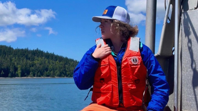 Devynn sits on a boat with her head turned to the side, looking off into the distance. She is wearing a life vest, rubber field pants, and other cold weather clothes.