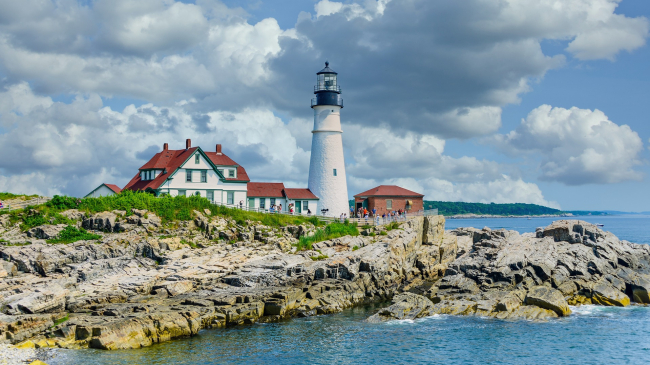 Photo of the Portland Head Light in Cape Elizabeth, ME. The white lighthouse with a black top stands on a small promontory next to a white house with green trim and a red roof.