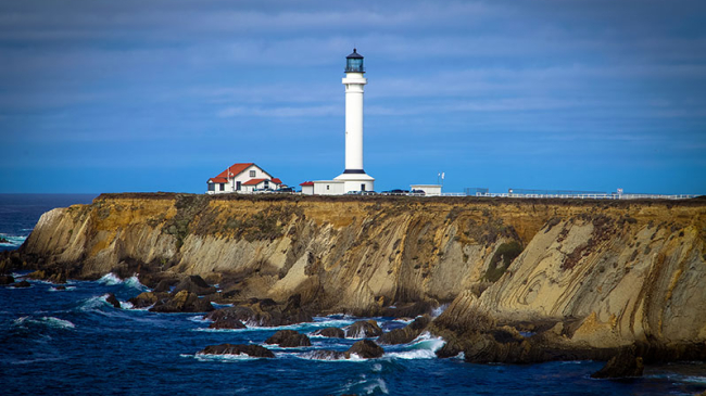 Photo of Point Arena Lighthouse in Point Arena, CA stands overlooking Greater Farallones national marine sanctuary.The tall, white lighthouse with a black light structure stands on a bluff that stretches out into the ocean. Next to the lighthouse is a low white house with a red roof.
