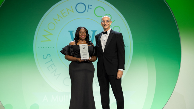 Natasha and the conference host, Eric S. Musser, stand side-by-side in black-tie attire as Natasha accepts a placard as her award. They stand in front of a screen that is projecting the Women of Color in STEM logo.