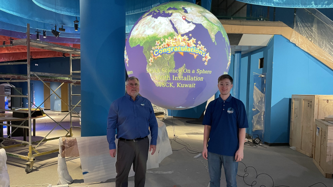 Two men stand in front of a Science On a Sphere globe that displays a world map and the word ‘congratulations’. The globe hangs in an unfinished museum gallery, with parts and boxes on the floor around the site.