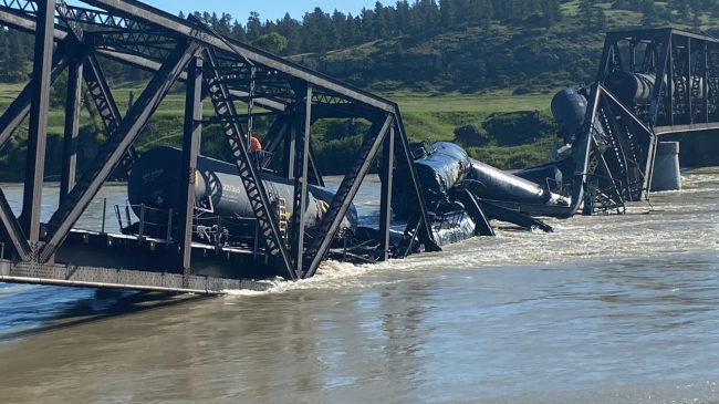 Rail cars, impacted by a train derailing on a bridge over the Yellowstone River in Montana, are observed semi-submerged between the damaged bridge and the river.