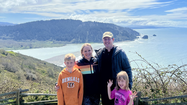 Two adults wearing jackets are flanked by their two children at the Klamath River Overlook. Behind them is the mouth of the river where it meets the Pacific Ocean. On either side of the river are two hills covered in shrubs and pine trees.