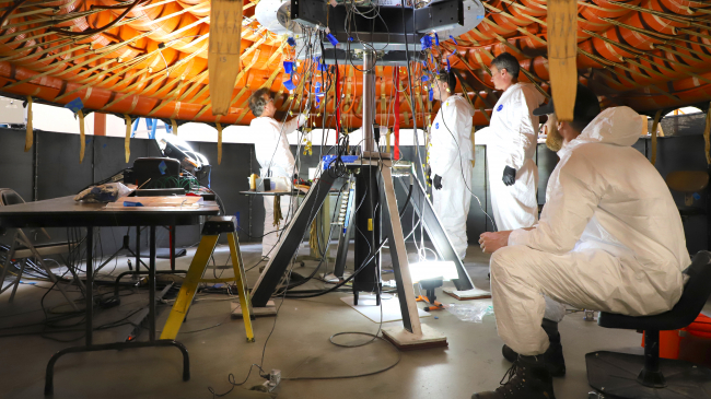 Engineers prepare for the flexible heat shield installation on the inflatable structure. The view is from bottom side, heat shield is on top. The gold straps hanging from the black triangles will be attached to the inflatable structure strapping and tensioned. 