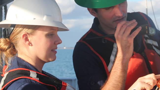 Two people working on a ship wearing protective headgear and high visibility vests.